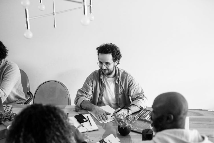 Black and white photo of a man with glasses sitting at a table with papers, surrounded by three other people, having a meeting or discussion in a modern office setting.