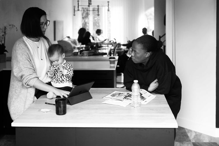 Two adults stand at a kitchen island; one holds a baby and uses a tablet, while the other leans on the counter next to an open magazine, a water bottle, and a mug. The scene is bright and modern.