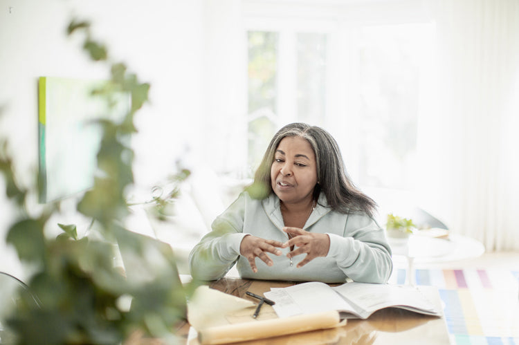 A woman with long gray hair sits at a table indoors, gesturing with her hands. She is surrounded by open books and papers, with a plant in the foreground and sunlight streaming through large windows behind her.