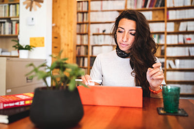 A woman with long brown hair sits at a wooden table using a tablet and stylus. She wears a white top and a scarf, with books and a potted plant on the table. Shelves filled with books are in the background.