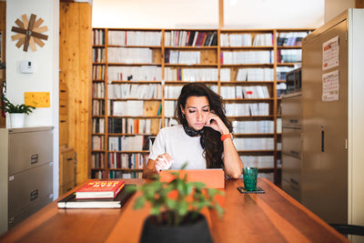 A woman sits at a wooden table in a library or office, reading a tablet. Books and a drink are on the table, and shelves filled with books line the background. A plant is in the foreground.