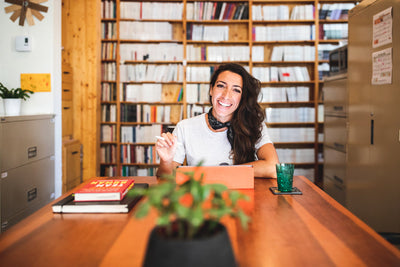 A woman with long brown hair, smiling and sitting at a wooden table in an office or library, with bookshelves behind her, a plant in the foreground, and a glass of water and books on the table.