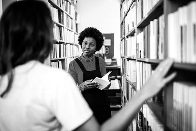 Two people stand between bookshelves in a library. One person, holding an open book and wearing a striped shirt under overalls, looks at the other person, who is seen from behind. The image is in black and white.