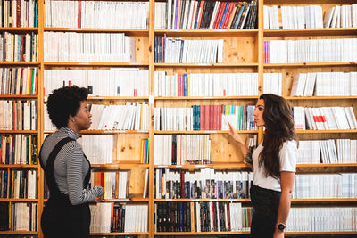 Two women stand facing each other in front of a large bookshelf filled with books, having a conversation in a brightly lit library or bookstore.
