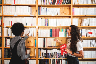 Two women stand in front of bookshelves filled with books. One woman, holding a red book, talks to the other, who is reaching for a book on the shelf. They appear to be having a discussion in a library or bookstore.