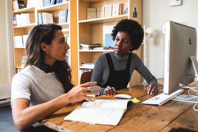 Two women sit at a wooden desk in an office, one speaking while gesturing with her hand, the other listening attentively. An open notebook and a desktop computer are on the table, with bookshelves in the background.