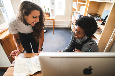 Two women smiling and talking together in an office, one standing while holding a pencil and notebook, the other seated at a desk with an open book and an Apple computer in front of them.