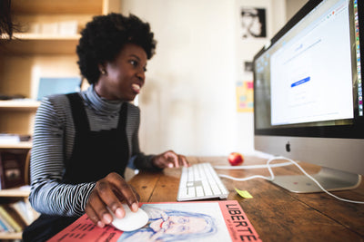 A woman smiles while working at a desktop computer in a cozy home office, using a mouse and keyboard. There are bookshelves and a colorful poster on the desk.