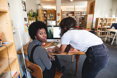 Two women work together in a bright office space; one sits at a desk looking at the camera while the other leans over the desk, writing on a piece of paper next to a computer monitor.