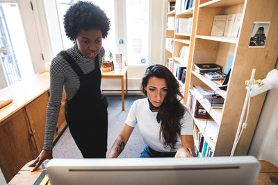 Two women work together in a bright office, one standing and one sitting at a desk using a computer, surrounded by bookshelves and natural light from a nearby window.