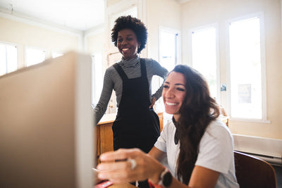Two women smiling and looking at a computer screen together in a bright, sunlit room. One woman is standing while the other is seated and using the keyboard.