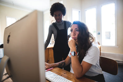 Two women work together at a computer in a bright office. One woman sits and focuses on the screen, while the other stands beside her, both appearing engaged and thoughtful.