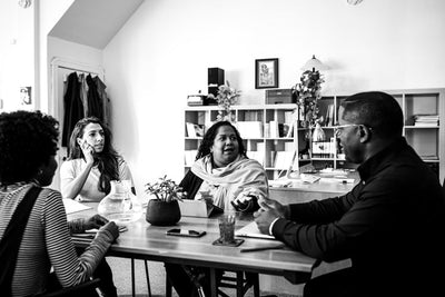 A black and white photo of four people having a discussion around a table in an office. Papers, notebooks, a plant, and a glass pitcher are on the table, and shelves with books and decor are in the background.