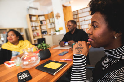 Three people sit around a wooden table in a cozy office, engaged in conversation. In the foreground, a woman with I CAN I WILL I DO written on her hand looks thoughtful. Books and a plant are on the table.