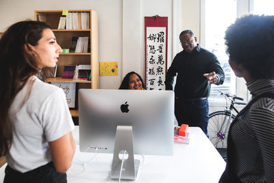 Four people gather around a desk with an iMac computer in a bright office. One person is seated and smiling, while the others stand and engage in conversation. Shelves, a bicycle, and wall art are visible in the background.