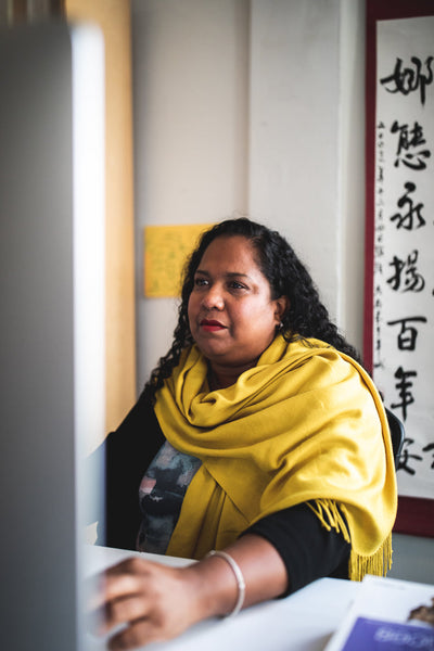 A woman with curly dark hair, wearing a yellow shawl, sits at a desk and looks at a computer screen. There is a scroll with Chinese characters hanging on the wall behind her.