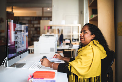 A woman with curly hair, wearing a yellow shawl, sits at a desk using a large desktop computer in a bright office space with shelves and a printer in the background.