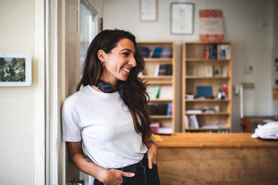 A woman with long dark hair, wearing a white t-shirt and a dark scarf, smiles as she leans against a doorway in a cozy, well-lit room with wooden bookshelves in the background.