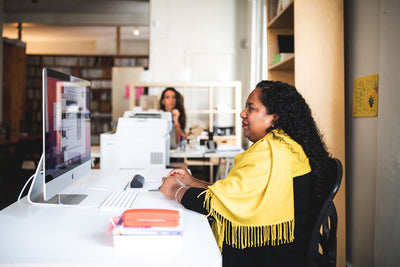 A woman wearing a yellow shawl sits at a desk working on a computer in a bright, modern office. Another person is visible in the background, also working at a desk.