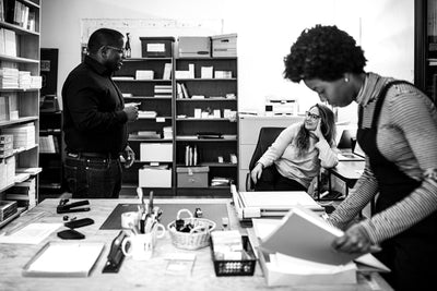 Three people work in an office with shelves of books and supplies. One man stands talking, a woman sits and listens, and another woman looks through papers at a desk in the foreground. The image is in black and white.