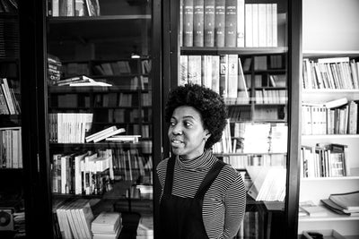 Black and white photo of a woman with curly hair, wearing a striped turtleneck and overalls, standing and smiling in front of bookshelves filled with books.