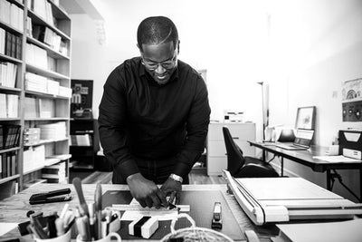 A man wearing glasses and a dark shirt uses a ruler to measure paper at a desk in an office filled with books, shelves, and office supplies. The atmosphere is bright and organized.