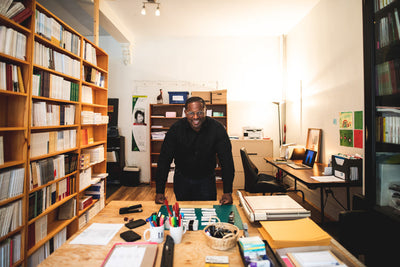 A person stands smiling behind a desk in a bright, organized office filled with shelves of books and various office supplies, creating a welcoming and productive atmosphere.