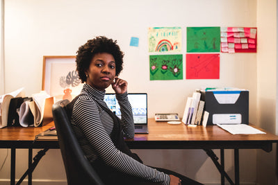 A woman sits at a desk in a home office, looking at the camera. Behind her are colorful drawings on the wall, office supplies, and a laptop. The scene is well-lit and organized.