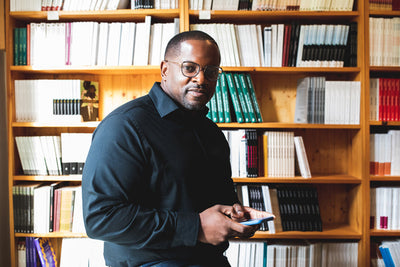 A man wearing glasses and a black shirt stands in front of bookshelves filled with books, holding a smartphone and looking at the camera.
