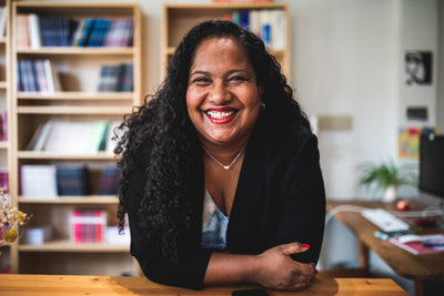 A woman with long curly hair smiles warmly while leaning on a wooden desk. She wears a black blazer and stands in a cozy, brightly lit office with bookshelves and plants in the background.