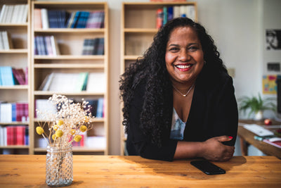 A woman with curly hair and a bright smile leans on a wooden table in a cozy room with bookshelves in the background; a vase with dried flowers and a smartphone sit on the table.