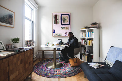 A person sits at a round table in a bright, cozy room working on a laptop. The space features bookshelves, a colorful rug, modern art on the wall, and natural light streaming through a large window.