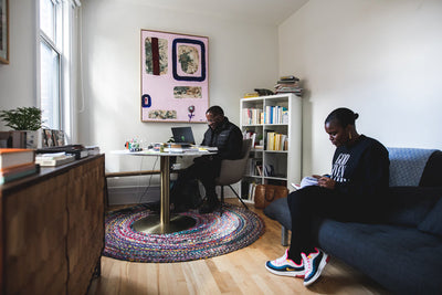Two people work in a cozy, sunlit room; one sits at a table with a laptop, another reads on a sofa. Shelves of books, colorful rug, and modern art decorate the space.