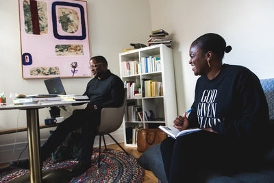 Two people sit in a cozy room: one at a desk with a laptop, the other on a couch smiling and writing in a notebook. Bookshelves and abstract artwork decorate the background.