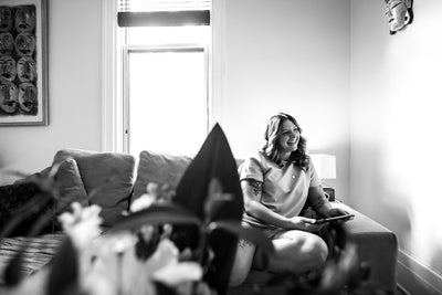 A woman sits on a couch by a window, smiling and holding a tablet. The room is bright, with light streaming in, and plants are visible in the foreground. A piece of art hangs on the wall behind her. Black and white photo.