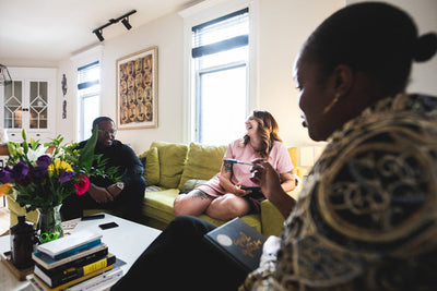Three people sit and chat in a bright living room with light walls, a green couch, books, and flowers on the table. Two people are on the couch, smiling, while the third sits in the foreground, holding a book.