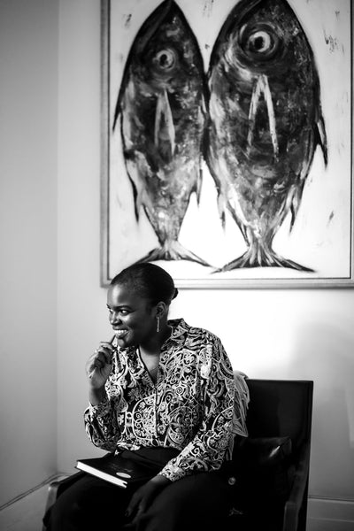 A woman sits and smiles while holding a pen, with a notebook on her lap. Behind her hangs a painting of two large fish. The image is in black and white.