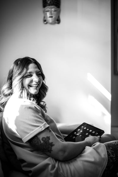 A woman with wavy hair smiles while sitting on a couch, holding a tablet. Sunlight streams through the window, illuminating her face. A decorative mask hangs on the wall behind her. The image is in black and white.