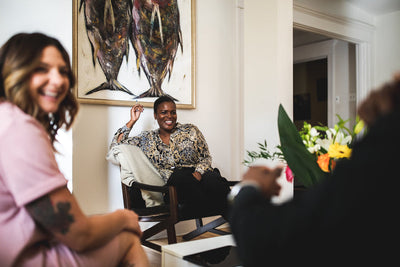 Three people sit and smile in a brightly lit living room with a large fish painting on the wall and a bouquet of flowers on the table in the foreground.