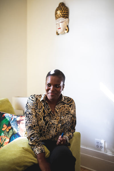 A person with short hair, wearing a patterned shirt, sits on a green couch in a well-lit room. A colorful pillow and a Buddha head wall decoration are visible in the background.