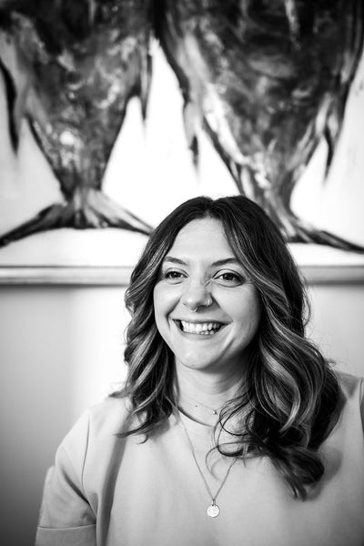 A woman with wavy hair smiles, wearing a light top and a necklace with a round pendant. The background features an abstract black-and-white painting. The image is in black and white.