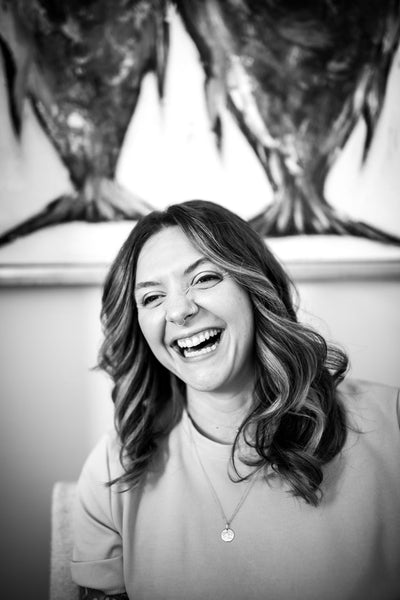 A woman with wavy hair smiles and laughs while sitting indoors. She wears a light shirt, a nose ring, and a necklace. The background features an abstract painting. The photo is in black and white.