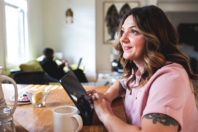 A woman with wavy brown hair and a pink top sits at a wooden table, smiling and holding a tablet. A coffee cup, a glass of water, and a lemon wedge are on the table. Another person sits in the background by the window.
