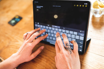 Close-up of tattooed hands typing on a tablet with an on-screen keyboard, placed on a wooden table. A drink and a card are visible in the background.