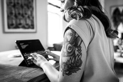 A woman with a detailed deer and floral tattoo on her upper arm types on a tablet at a wooden table in a bright room. The image is in black and white.