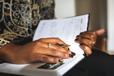 Close-up of a person‚Äôs hands holding a pen and writing in a planner or notebook. The person has dark nail polish and is wearing a gold ring and a patterned, ornate sleeve.