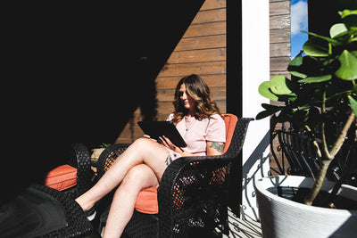 A woman sits on a patio chair outside, holding a tablet. She is wearing a pink dress and is partially in sunlight. There is a large potted plant beside her and a wooden wall in the background.