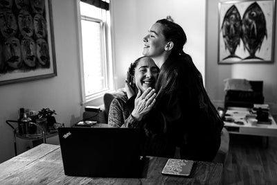 A woman sitting at a table with a laptop smiles as a younger woman hugs her from behind in a warmly lit living room. Both appear happy and affectionate.