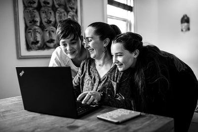 A woman and two children smile and look at the screen of a laptop together at a wooden table in a bright room. A painting of faces hangs on the wall in the background.