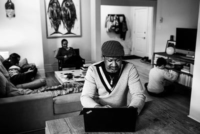 A man sits at a table using a laptop in a living room, with three children in the background engaged in different activities. The scene is in black and white, giving it a candid, everyday atmosphere.